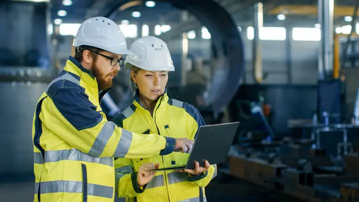 Two colleagues in hi-vis and hard hats reviewing a procedure on a laptop