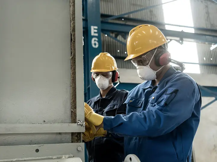Two workers in hard hats, masks and gloves working at a piece of industrial equipment